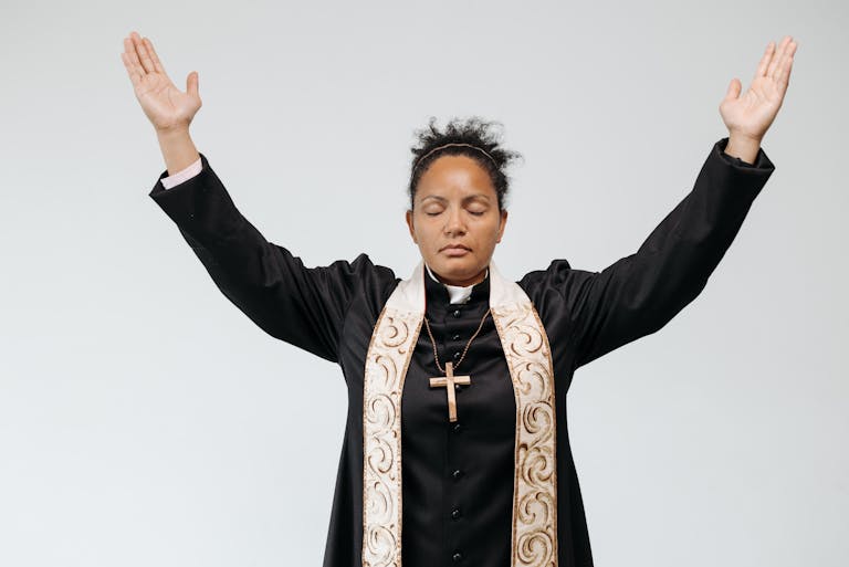 How to hear God for your business and there is a female priest hear from God for your business, is in black robe praying with arms raised and eyes closed against a white studio background.