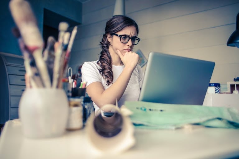 First 90 days Young woman with glasses deeply focused on a laptop surrounded by art supplies in a home office.