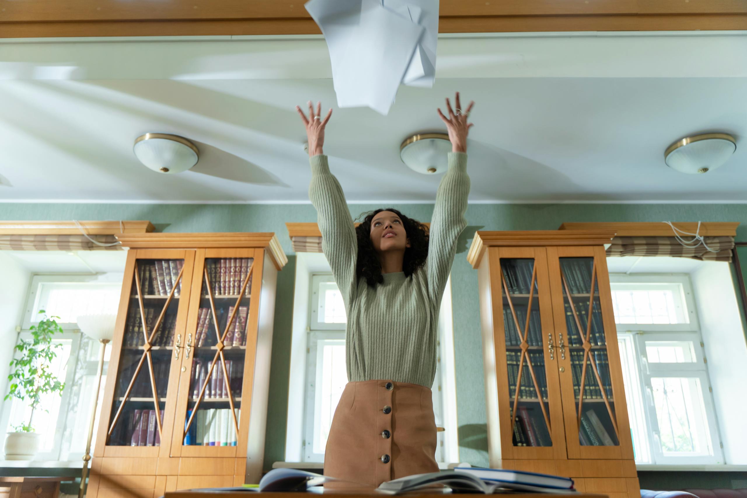 Long-term success of A young woman throws papers in the air in a library, expressing joy and relief.
