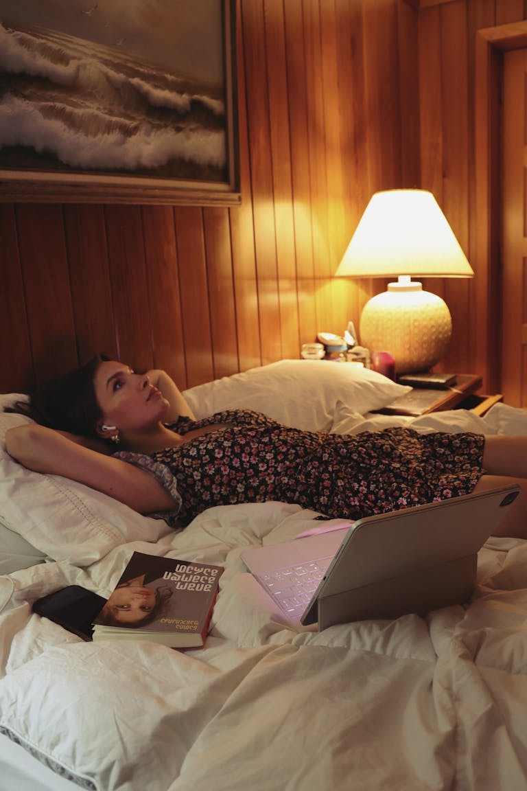 solutions to overthinking. A woman in a floral dress relaxes on a bed in a cozy room, with a book and a laptop nearby.